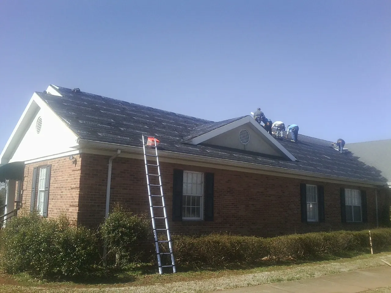 Crew tearing shingles off a brick ranch roof with a ladder set — Emergency Roof Repair work by our crew in Wray