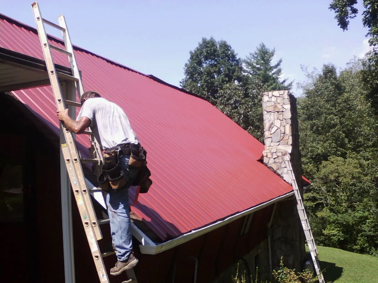 Roofer on a ladder beside a red metal roof and stone chimney — Soffit & Fascia Repair by Roofing Brothers in Wray